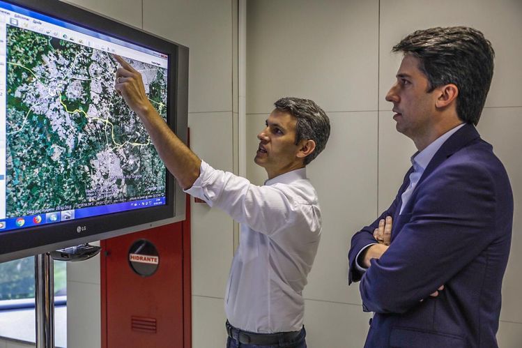 Gilson Santos apontando para uma televisão onde aparece o mapa da região metropolitana de Curitiba com o secretário de planejamento em pé olhando para a televisão. 